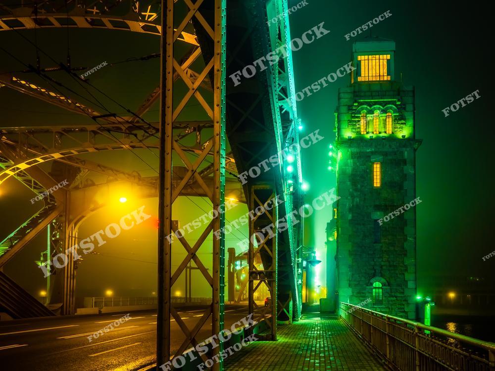 На фотографии изображён Большеохтинский мост, освещённый мягкой зелёной подсветкой, подчёркивающей его ажурные конструкции и массивную башню. Вечерний туман придаёт снимку загадочность и уют. Это место является одним из символов инженерного искусства Санкт-Петербурга, соединяя берега реки Невы.